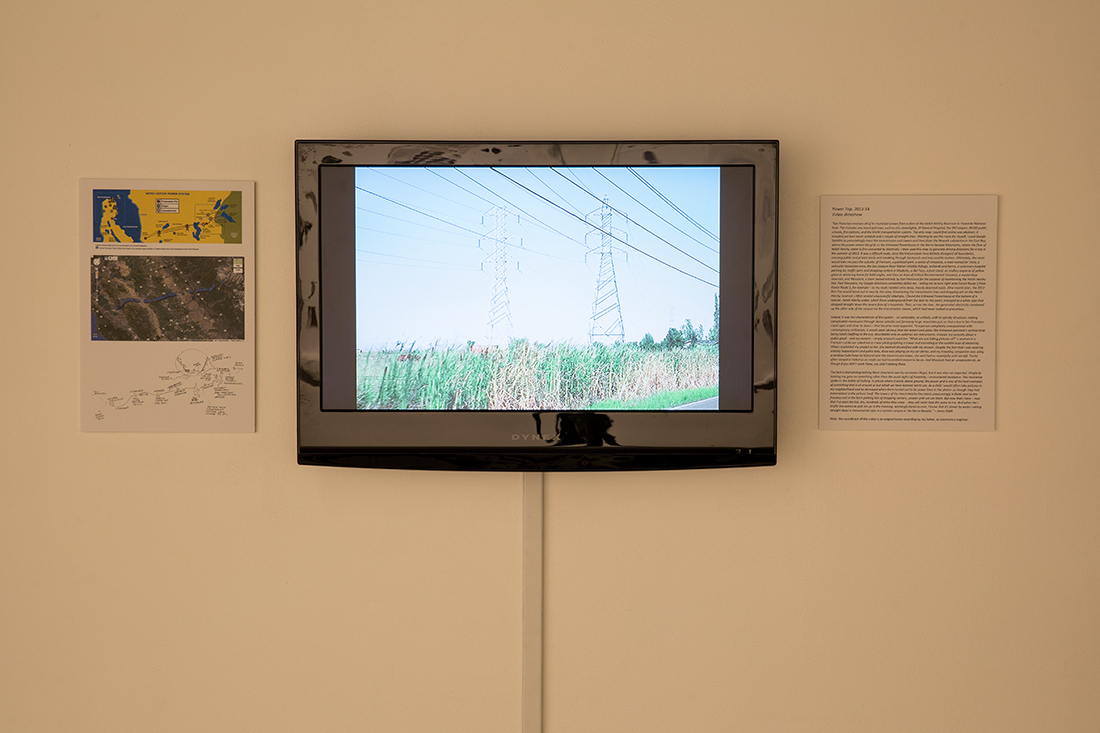 A wall-mounted screen showing a video of the transmission towers, with maps and information on one side, and a short essay on the other