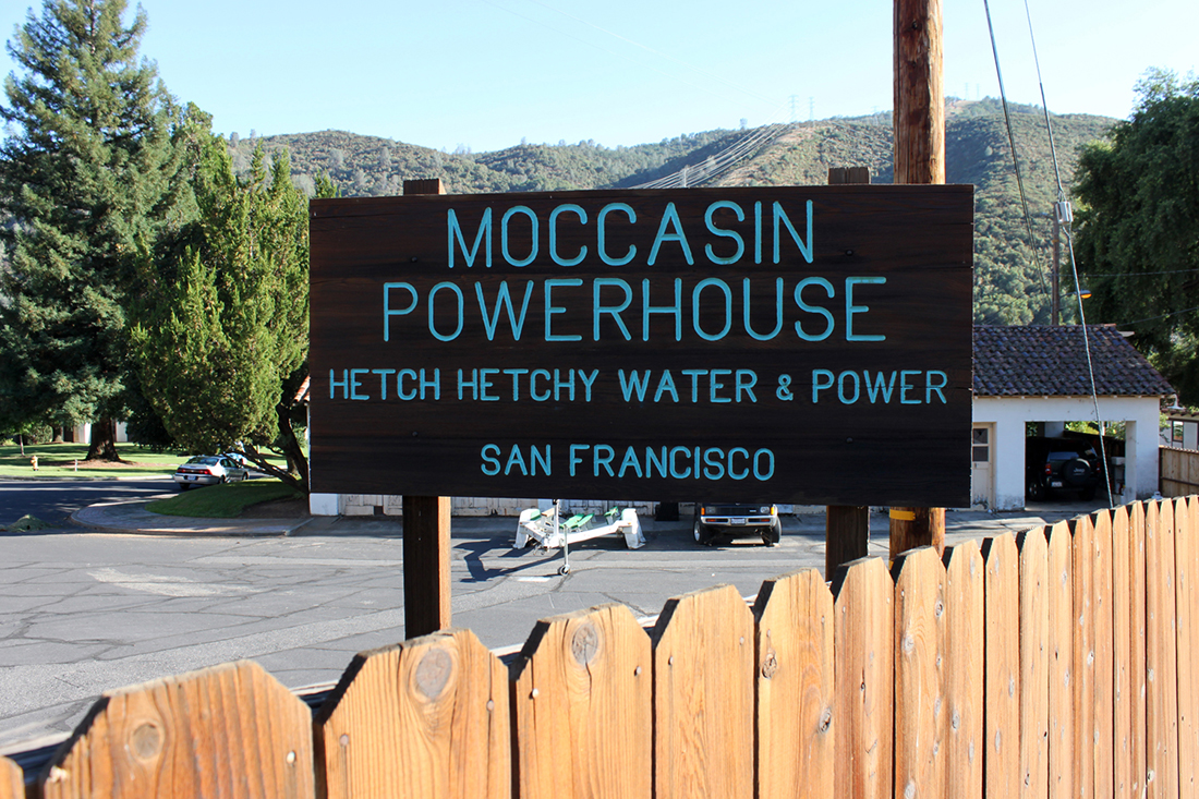 sign that says: Moccasin Powerhouse, Hetch Hetchy Water & Power, San Francisco, with power lines crossing forested mountains in the background