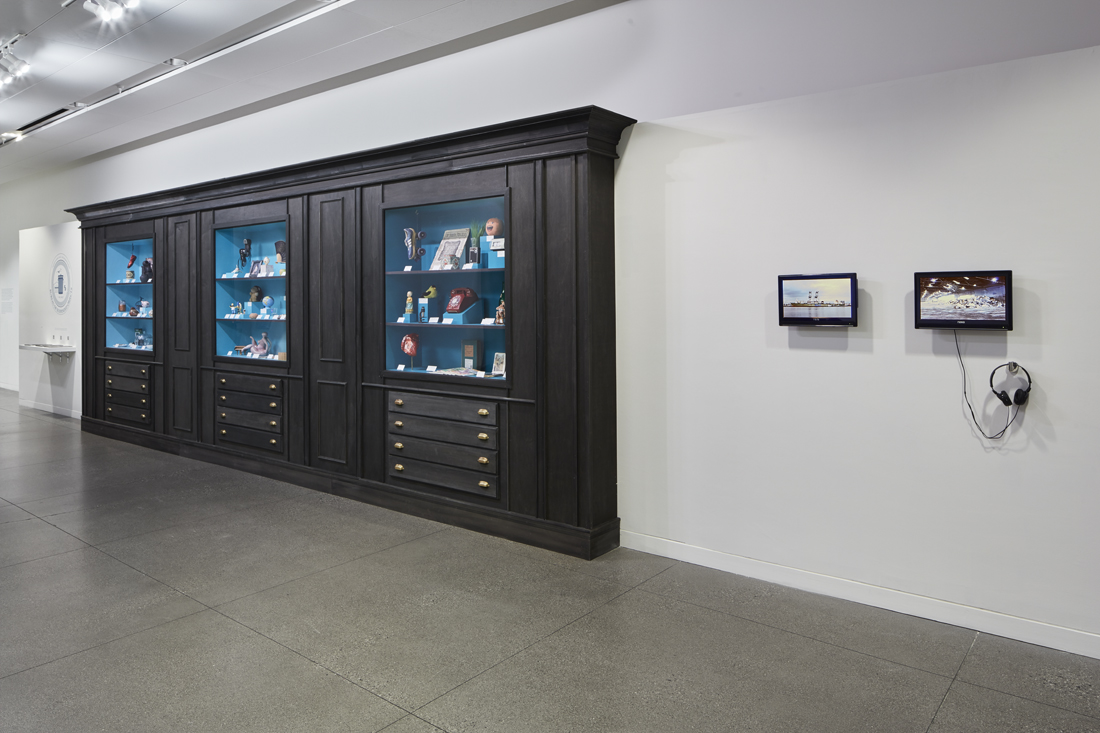 An overall view of the installation including the wooden display case, the shelf of books, and two wall-mounted video monitors showing footage of the Oakland port and the dump