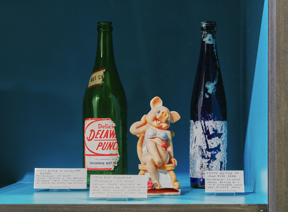 a closeup of the rightmost display, showing an empty bottle of Delaware Punch, a ceramic figurine of a pig in a bikini, and an empty bottle of sake