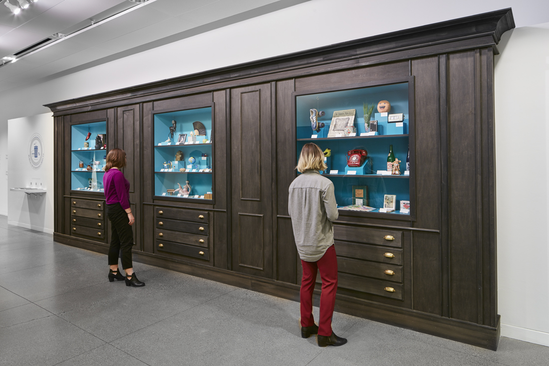 Two people in a museum hallway looking at wooden a display case, which has three separate display sections, each containing a handful of decorative and household objects