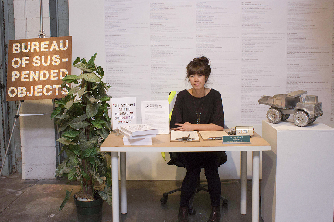Jenny Odell sitting at a small desk with the book, The Archive of the Bureau of Suspended Objects, and other information. On either side are a large sign for the Bureau of Suspended Objects made from found materials, a found artificial office plant, and on a pedestal, a found silver-painted Tonka truck that is part of the archive.