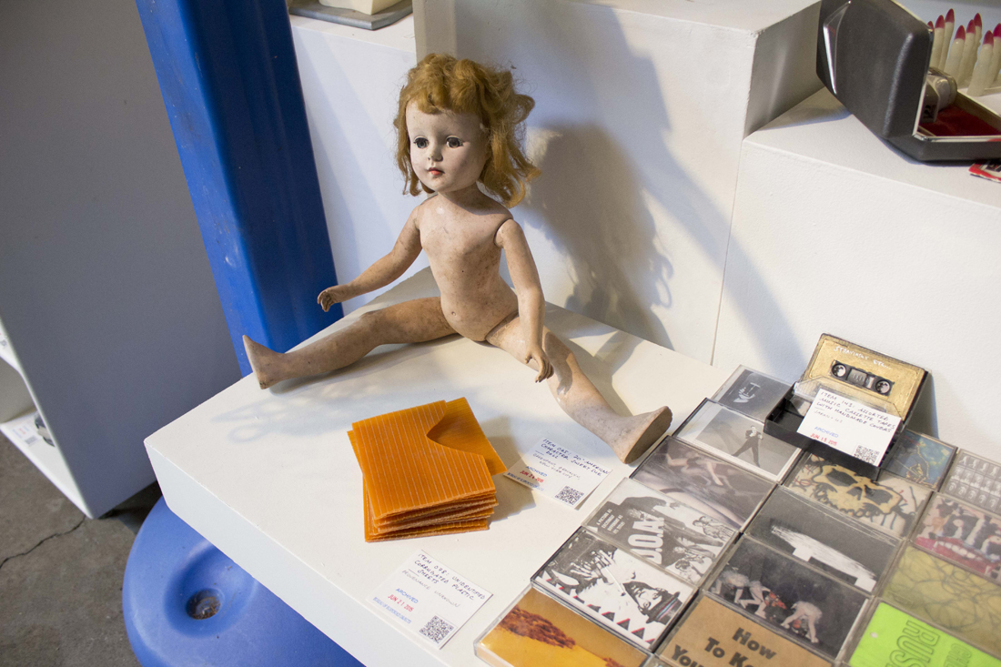 A shelf showing an old plastic doll, squares of plastic orange material, and cassette tapes, each with identifying tags