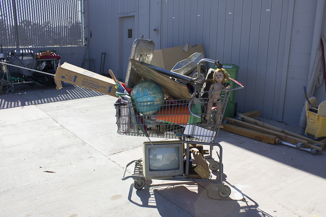 Outdoor shot of a shopping cart filled with stuff: a doll, globe, rainbow umbrella, television set, and Tonka truck