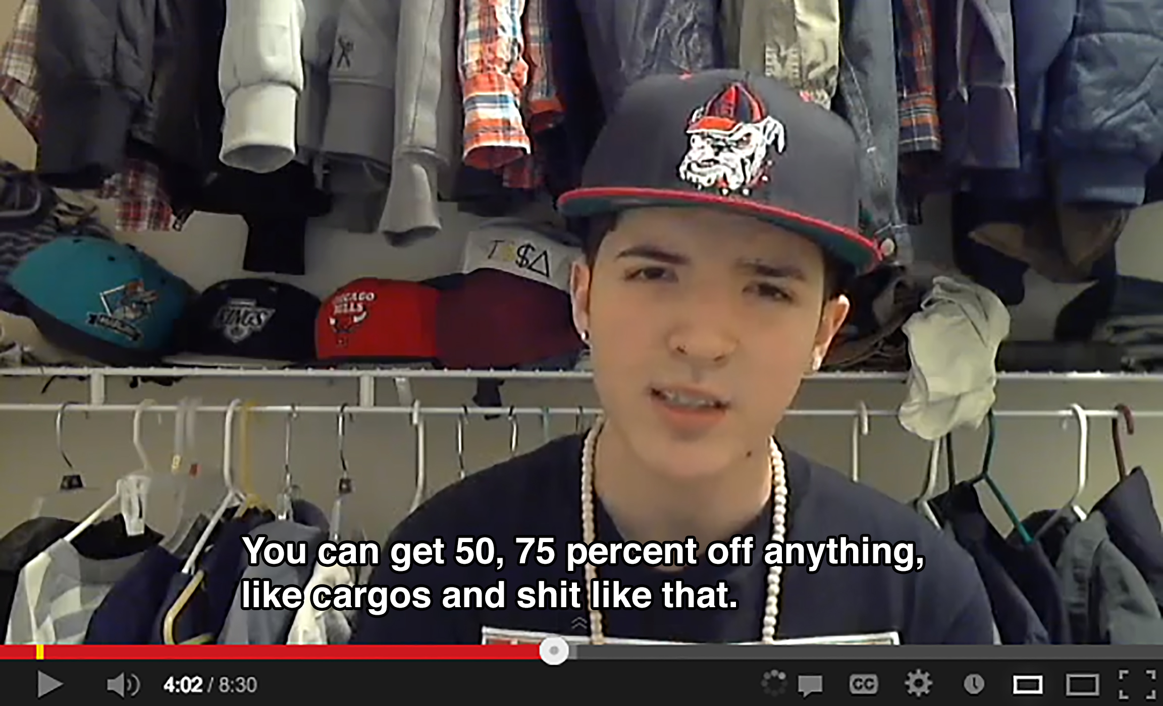 A teenager wearing a hat with a bulldog on it stands in his closet, which has many hats and jackets, and says: You can get 50, 75 percent off anything, like cargos and shit like that.