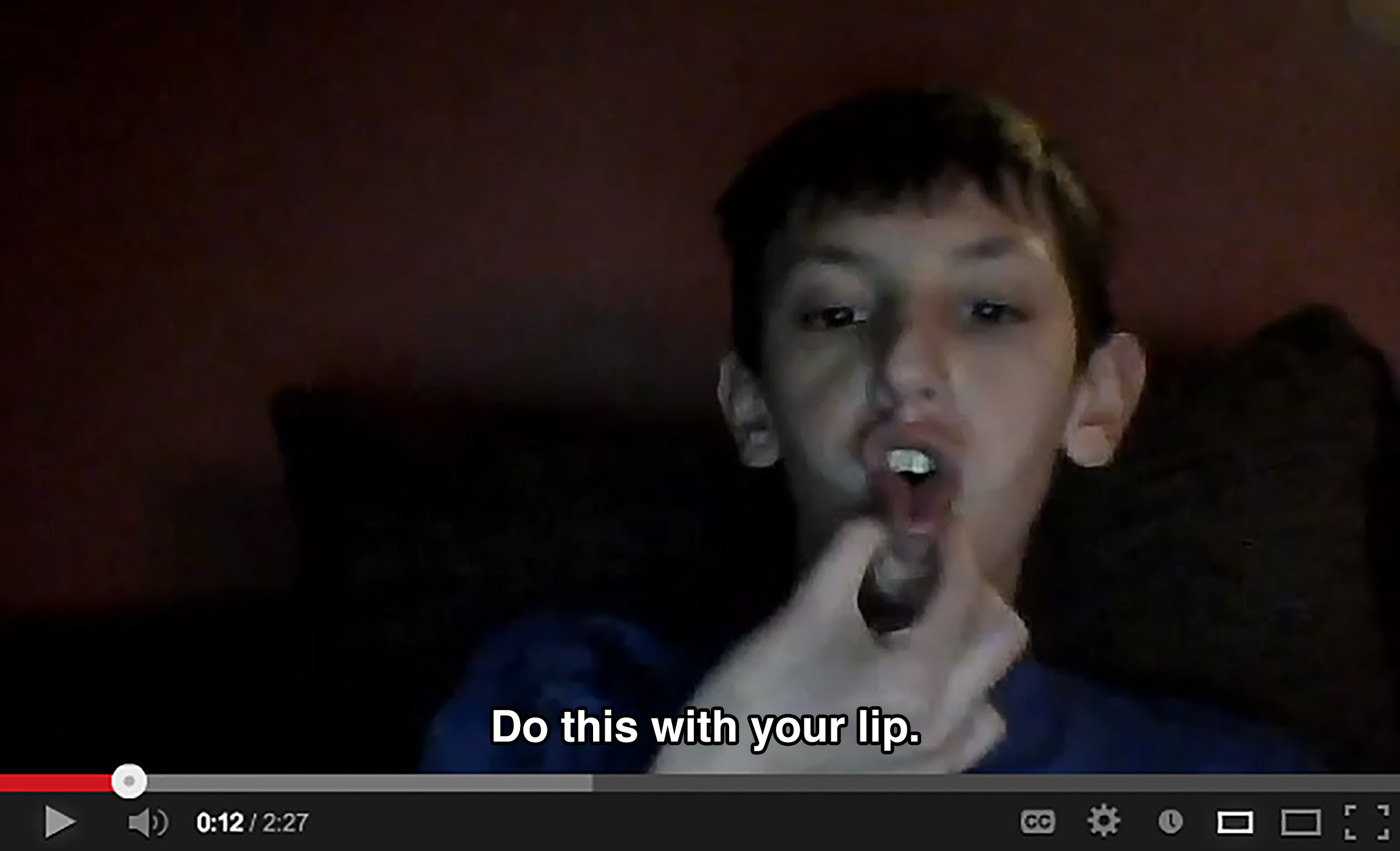 A boy in a dark room pulls his bottom lip downward and squishes it together, saying: Do this with your lip.