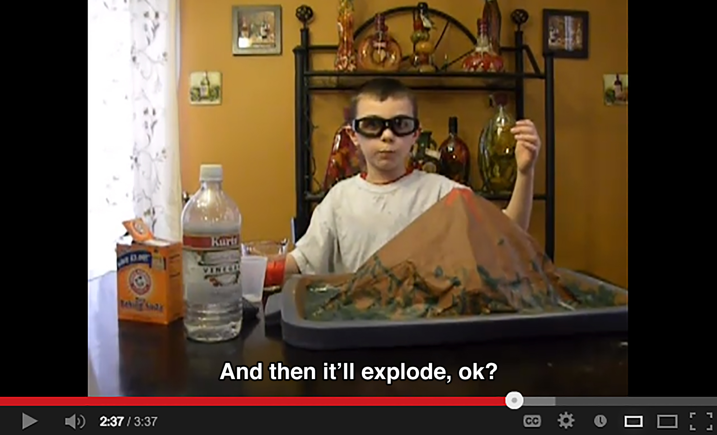 A young boy wearing goggles in a living room stands behind a model of a volcano, a bottle of vinegar, and baking soda, and says: And then it'll explode, ok?