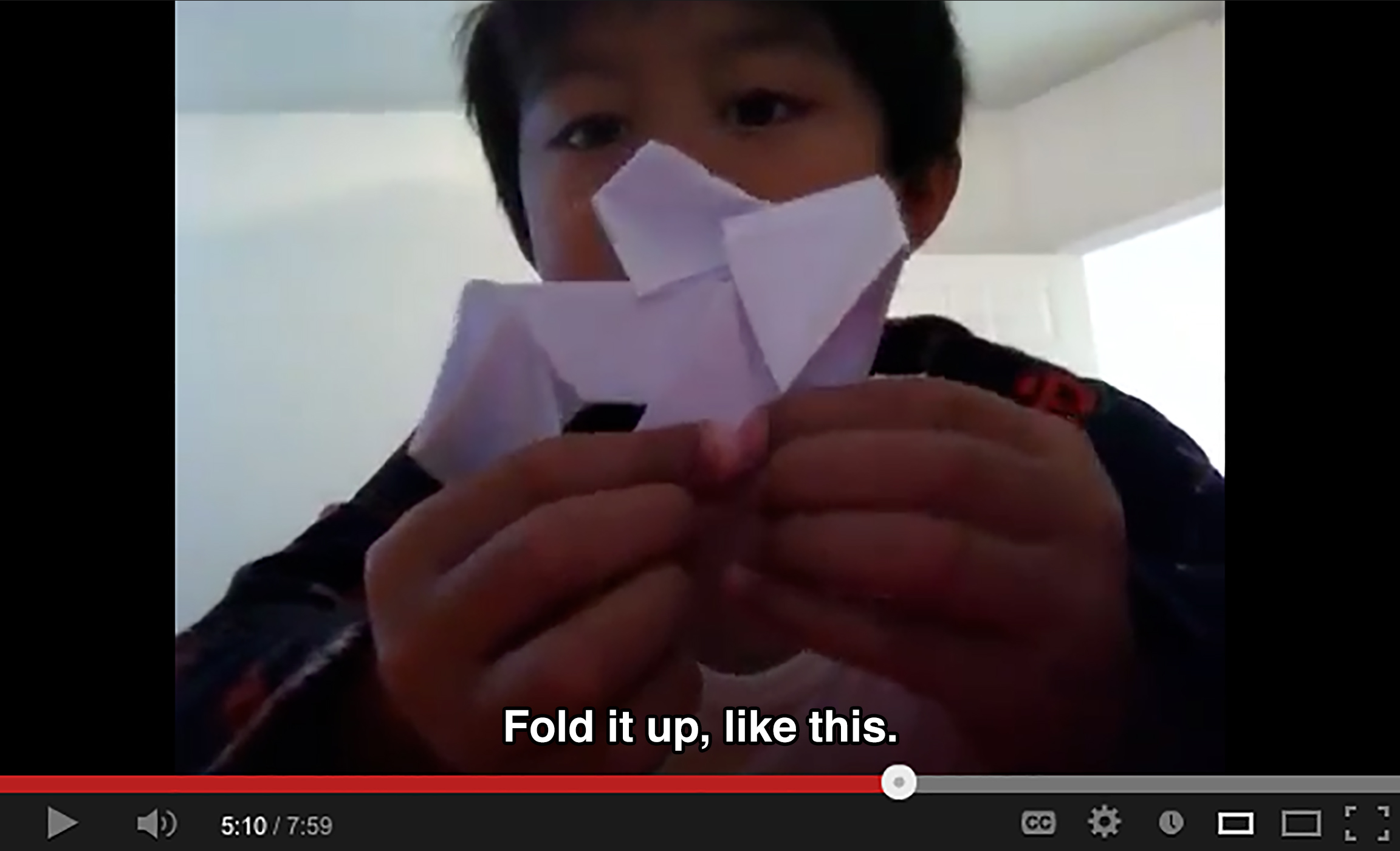 A young boy holds origami up to the camera and says: Fold it up, like this.
