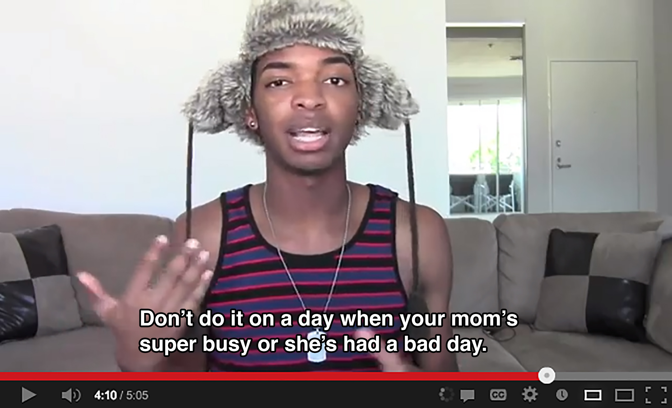 A teenager wearing a tank top and a trapper hat in a living room gestures with his hand and says: Don't do it on a day when your mom's super busy or she's had a bad day.