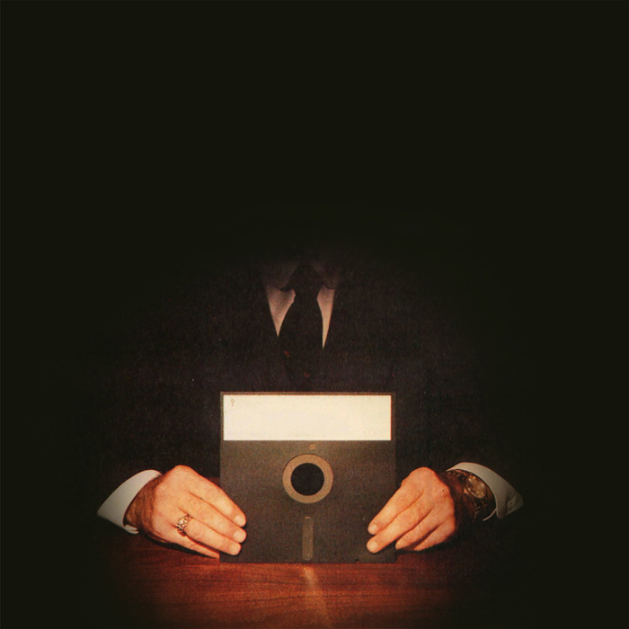 A formal businessman sitting at a table holding a floppy disk upright, with a spotlight on the disk so that his face is obscured by dark