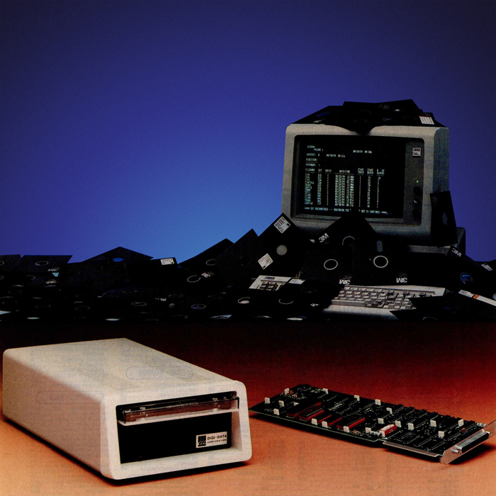 In the foreground, a disk drive and circuit board; in the background, a computer display covered in a messy pile of floppy disks