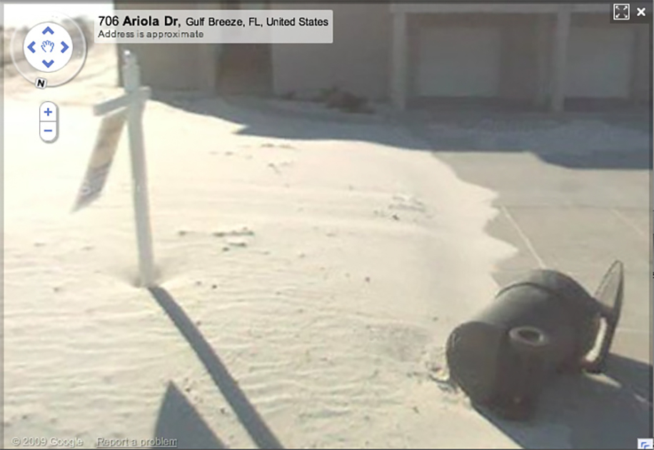 Screenshot of Google Street View of the front yard of a house in Florida, where the ground is covered entirely in sand, a For Sale sign blows in the wind, and a trash bin has been knocked over